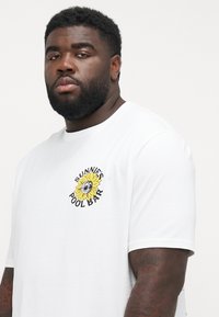 White t-shirt featuring a colorful sunflower graphic with sunglasses and the text "SUNNIE’S POOL BAR" in black. Short sleeves and crew neck design.
