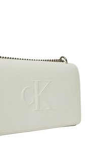Borsa a mano in pelle bianca con una superficie liscia, caratterizzata da un logo "ck" impresso. Dotata di una tracolla in catena argentata e chiusura con zip.