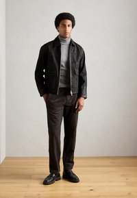 Giubbotto di pelle nera con due tasche frontali, maglione grigio a collo alto, pantaloni in velluto a coste marrone e mocassini neri. Texture lisce e design aderente.