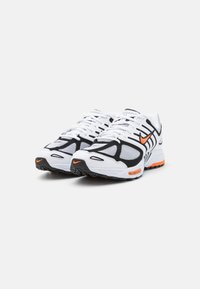 White athletic shoes with black and orange accents. Features mesh panels, a textured sole, and prominent brand logo near the heel.