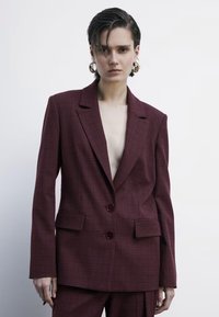 Blazer a quadri bordeaux con revers a punta, due bottoni e tasche frontali. Il tessuto ha una finitura texturizzata, che conferisce una silhouette strutturata.
