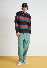 Stripet langermet skjorte i svart, grønn, oransje og blå. Løs passform grønn bukse med glidelåser på sidene. Lys blå sneakers.