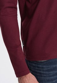 Camisa burdeos de manga larga con dobladillo redondeado, de material de algodón, etiqueta en la costura lateral y una textura suave, combinada con jeans azules.