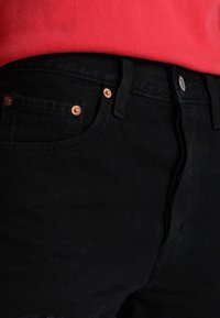 Jeans de mezclilla negra con herrajes en tono cobre en la cintura y los bolsillos, con una textura suave y un diseño ajustado.