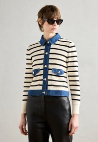Cardigan a righe nei toni crema e nero con colletto in denim e tasche. Presenta bottoni metallici e un taglio cropped. Abbinato a pantaloni neri.