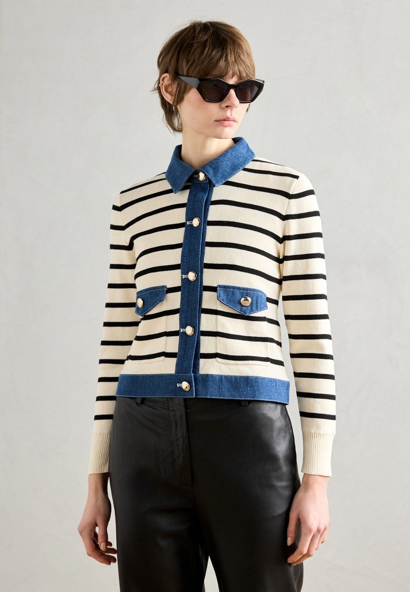 Cardigan a righe nei toni crema e nero con colletto in denim e tasche. Presenta bottoni metallici e un taglio cropped. Abbinato a pantaloni neri.