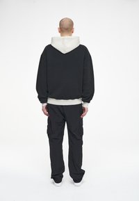 Uomo con testa rasata, che indossa un hoodie nero con cappuccio grigio, pantaloni cargo neri e sneakers bianche, in piedi di spalle su uno sfondo bianco.