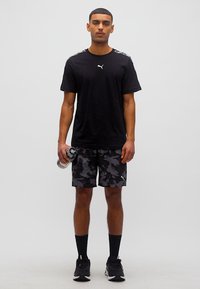 Νέος άνδρας που φοράει μαύρο T-shirt Puma και καμουφλάζ σορτς, κρατώντας ένα μπουκάλι νερό, στέκεται μπροστά σε λευκό φόντο.