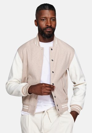 Man wearing beige and white varsity jacket over white shirt, fastening a button with one hand while the other rests in pocket.