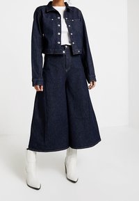 Tummansininen farkkutakki hopeisilla nappeilla, yhdistetty leveälahkeisiin tummansinisiin farkkuihin. Päällä valkoiset nilkkurit, joissa on klassinen muotoilu.