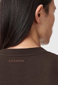 Osoba o długich ciemnych włosach ubrana w brązową koszulę z logo "ALLSAINTS" nadrukowanym przy dekolcie na plecach.