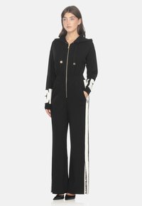 Tuta felpato nero con zip davanti, vita elastica e maniche lunghe. Accenti del logo bianco sulle maniche e sui lati, caratterizzato da una vestibilità ampia e gambe larghe.