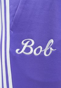 Μωβ ύφασμα με λευκό κεντημένο καλλιγραφικό κείμενο «Bob» και κάθετες λευκές ρίγες στην αριστερή πλευρά.