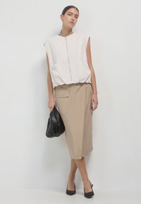 Femme portant un haut beige sans manches à fermeture éclair, une jupe beige mi-longue avec poche, tenant une pochette noire et des talons aiguilles noirs, sur fond blanc.