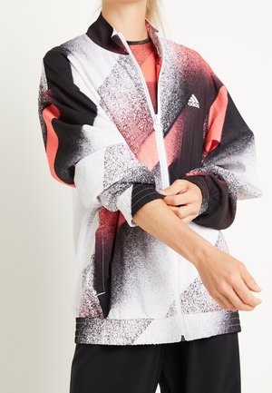 Person, der justerer ærmet på en hvid, sort og koralfarvet Adidas lynlåstrøje med abstrakt geometrisk mønster, stående mod en lys baggrund.