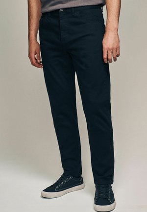 Marineblauwe katoenen broek met een rechte pijp, voorzien van een knoopsluiting, zijzakken en gedragen met donkere sneakers.