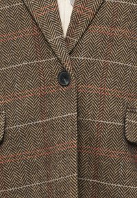 Brauner Fischgrätmuster-Blazer mit roten und blauen Akzenten, ausgestattet mit einem einzelnen schwarzen Knopf und zwei Fronttaschen. Strukturierter Wollstoff.