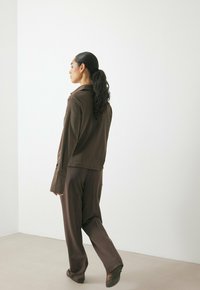 Bruin gemêleerd overhemd met kraag, gecombineerd met losse bruine broek en platte schoenen, gezien vanuit de achterkant. Eenvoudig en minimalistisch ontwerp.