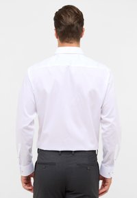 Camicia bianca con colletto a punta, texture liscia e maniche lunghe. Presenta dettagli con bottoni sui polsini. Indossata con pantaloni eleganti grigi.