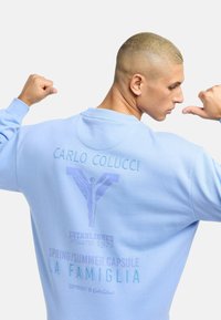 Svetlomodrá mikina s grafickým potlačou. Obsahuje logo "Carlo Colucci", postavu podobnú anjelovi a textové detaily v kontrastných odtieňoch.