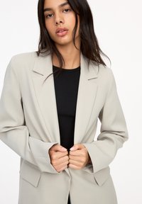 Hellgrauer Blazer aus glattem Stoff, mit lockerer Passform, Reverskragen und zwei vorderen Taschen. Getragen über einem schwarzen Oberteil.
