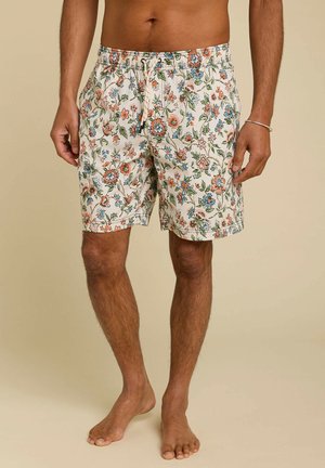 Personne torse nu portant un short blanc à motifs floraux avec des fleurs orange, bleues et vertes, debout pieds nus sur un fond clair.