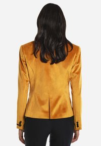 Blazer amarillo de terciopelo con un diseño ajustado, que presenta una abertura trasera y acentos de botones negros en las mangas. Textura suave y tersa.