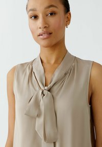 Beige ärmellose Bluse mit einer Bindekragen, leichtem Stoff, weicher Textur und subtilen Drapierungsdetails um den Kragen.