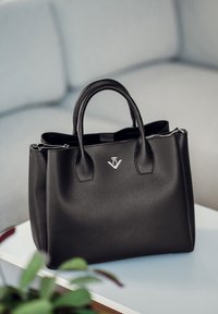 Schwarze Leder-Handtasche mit doppelten Griffen, Reißverschlussverschluss und einem Metall-Logo-Akzent auf der Vorderseite. Glatte Textur, strukturiertes Design.