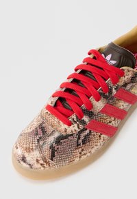 Sneakers con un motivo in similpelle di serpente beige e nero, dotati di lacci rossi e tre strisce rosse, con suola in gomma.