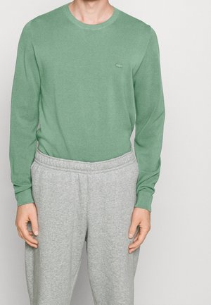 Homme portant une chemise verte texturée à manches longues avec un petit logo sur la poitrine et un pantalon de survêtement gris clair sur un fond uni.