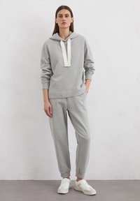 Grijze hoodie met witte trekkoorden, gecombineerd met grijze sweatpants met elastische boorden. Het model draagt witte sneakers, tegen een neutrale achtergrond.