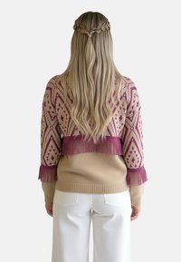 Dzianinowy sweter z geometrycznym wzorem w odcieniach różu i beżu, z frędzlami przy dole i rękawach, zestawiony z białymi spodniami.