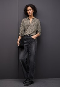 Camicia a maniche lunghe con stampa leopardata, jeans grigi a vita alta, stivali neri alla caviglia, mentre tiene una pochette nera, di fronte a un muro grigio scuro.