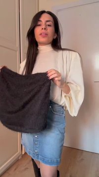 Γυναίκα με μακριά σκούρα μαλλιά, φορώντας μπλουζάκι εκρού και τζιν φούστα, κρατά ένα μαύρο, χνουδωτό ορθογώνιο ύφασμα σε ένα δωμάτιο.