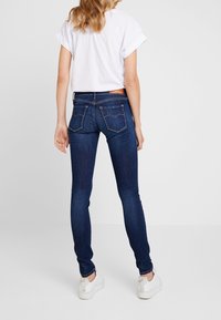 Vaqueros skinny de un azul oscuro con una cintura alta, que presentan dos bolsillos en la parte trasera y detalles de costura sutiles, combinados con una camiseta blanca de mangas enrolladas.