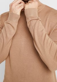 Beige langærmet sweater med høj krave, lavet af blødt strikmateriale. Har ribkantede ærmer og en glat overflade.