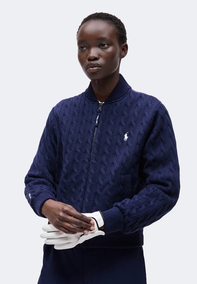 Personne portant un pull zippé en maille torsadée bleu marine avec un logo blanc, tenant des gants blancs sur un fond clair uni.