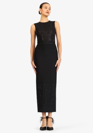 Frau in einem ärmellosen schwarzen Midikleid mit besetztem Oberteil und Saum, steht mit entspannten Händen, trägt schwarze Absätze mit Federdetails.
