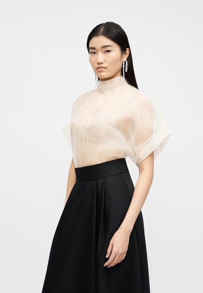 Femme aux cheveux noirs raides portant des boucles d'oreilles pendantes en cristal, un chemisier beige transparent à manches courtes et une jupe plissée noire taille haute, debout.