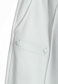 Tela de algodón blanca que muestra una textura suave, con un parche rectangular de cuero que cuenta con un herraje circular y un detalle de texto en relieve.