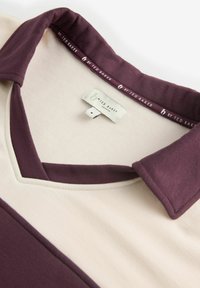V-kaelusega polo särk, millel on kahe tooniga disain kreemi ja tumepunases värvitoonis, valmistatud pehmest kangast, mille sildil on kirjas "By Ted Baker."