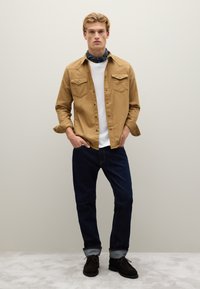 Lichtbruine button-up overhemd met twee borstzakken, witte t-shirt eronder, donkere spijkerbroek en donkere schoenen. Glad textuur, casual stijl.