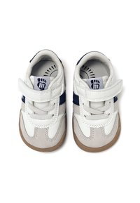 Zapatillas para niños en cuero blanco y marino con una correa de velcro, detalles en ante gris y una suela de goma marrón. El interior presenta un diseño de huella.