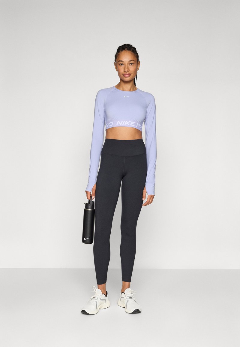 Top cropped de manga larga color púrpura claro con el texto "PRO", combinado con leggings negros de tiro alto y zapatillas blancas. Botella de agua negra sostenida en la mano.