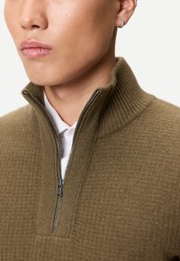 Nahaufnahme einer Person, die einen braunen, strukturierten Reißverschluss-Pullover über einem weißen Hemd mit Kragen trägt, mit einem kleinen silbernen Creolenohrring.