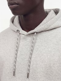 Gros plan d'une personne portant un sweat à capuche gris clair avec des cordons à motifs et des embouts métalliques, montrant le cou et le bas du visage.