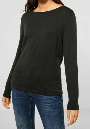 Frau trägt einen schlichten schwarzen Pullover mit langen Ärmeln und gerippten Bündchen sowie eine blaue Jeans, steht vor einem hellgrauen Hintergrund.