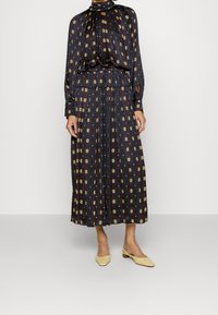 Maxime dressi tummasta kankaasta, värikkäällä ympyräkuviolla, korkealla kauluksella, pitkillä hihoilla ja kapealla vyötäröllä, yhdistettynä keltaisiin matalapohjaisiin kenkiin.