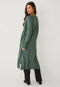 Cardigan lungo verde in un tessuto morbido con una vestibilità rilassata, caratterizzato da maniche lunghe, spacco laterale e polsini a costine, abbinato a pantaloni neri.